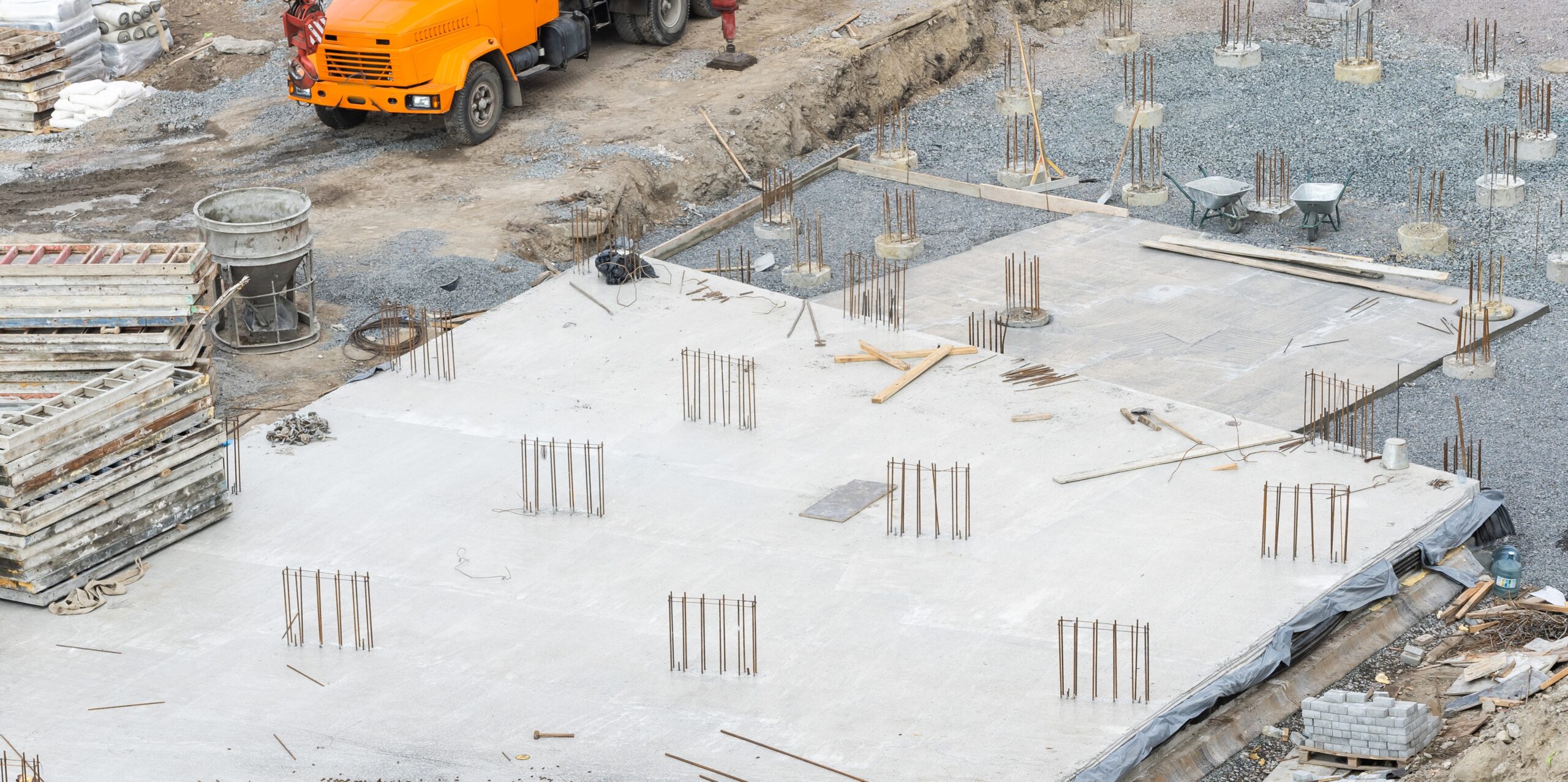 A construction site with concrete foundation, rebar protruding, materials stacked, and an orange truck in the background.